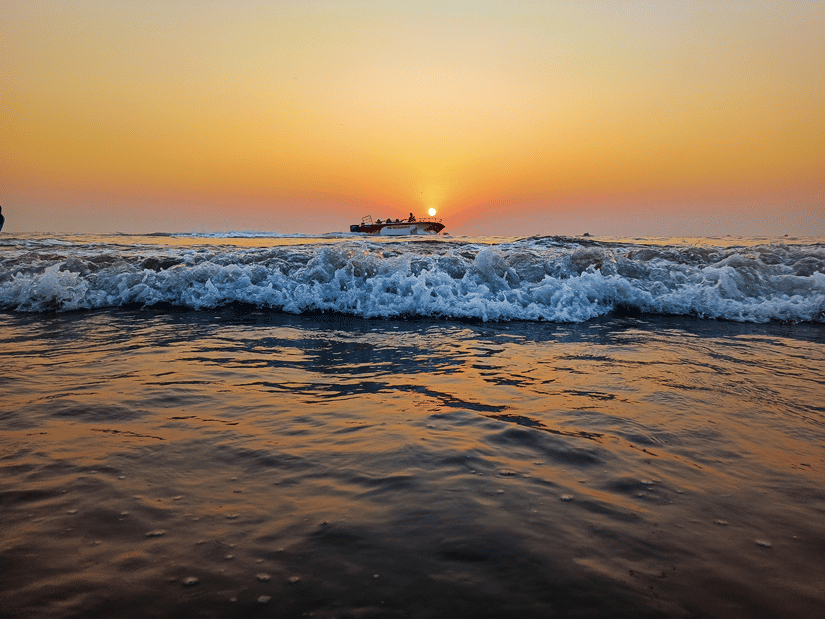Low-angle waves crashing onto the shore, with a lone boat silhouetted against a golden sun rising over the misty ocean horizon.