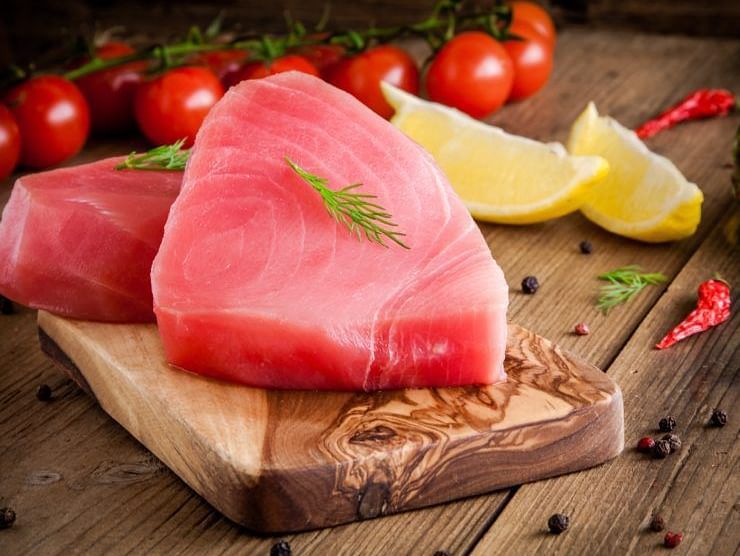 Fresh tuna steak on a wooden board, garnished with herbs and surrounded by lemon and tomatoes.