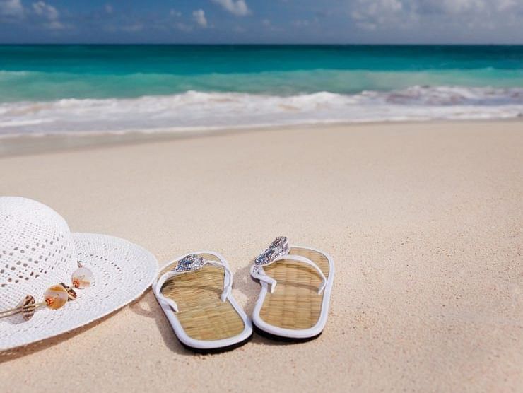 An image showing a white hat, slippers on the sand with ocean waves in the background.