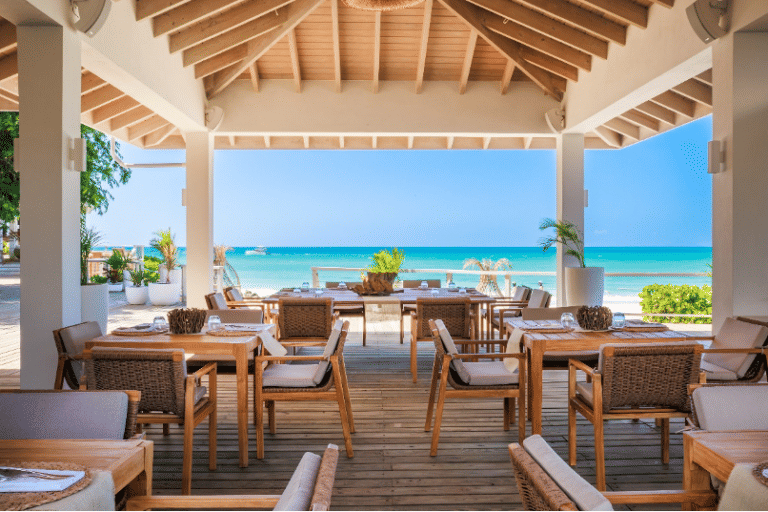 An open-air restaurant with wooden tables and chairs, offering panoramic views of a turquoise ocean and clear sky.