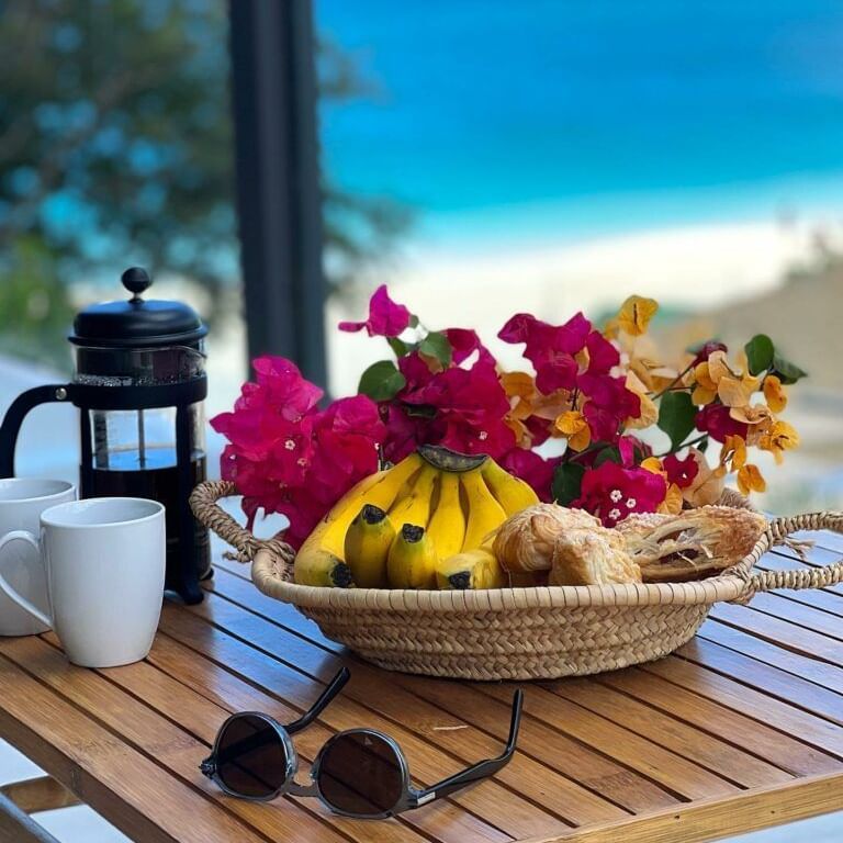 A wooden table with fresh fruit, pastries, coffee, and sunglasses, overlooking a vibrant blue ocean.