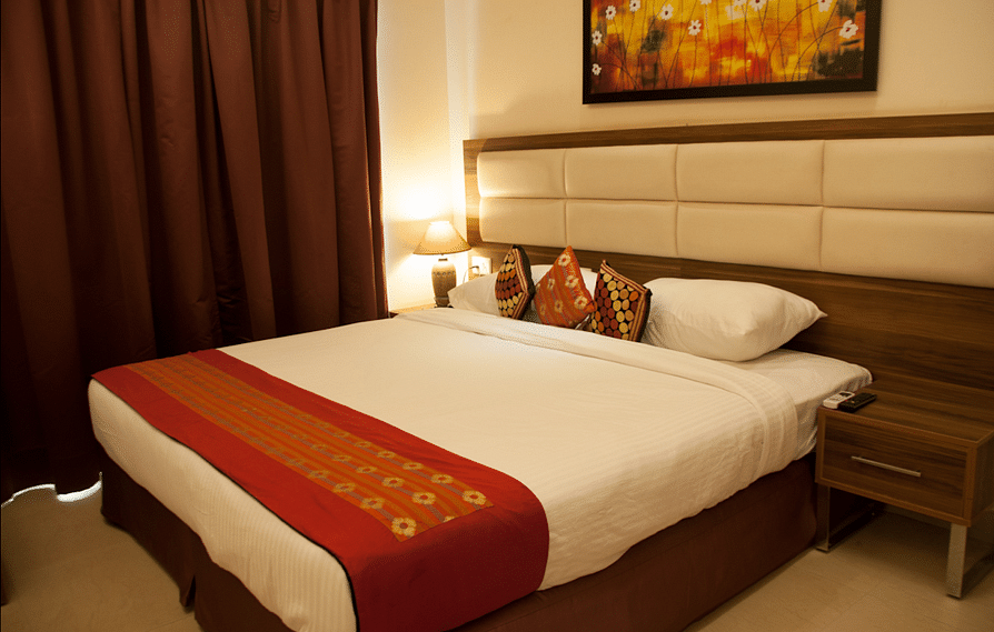 A neatly made hotel room bed with a decorative red runner and pillows, beside a nightstand and drawn curtains | Astra Hotels and Suites Electronic City