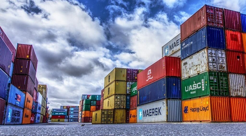 Stacked colourful shipping containers under a cloudy sky at a logistics yard.