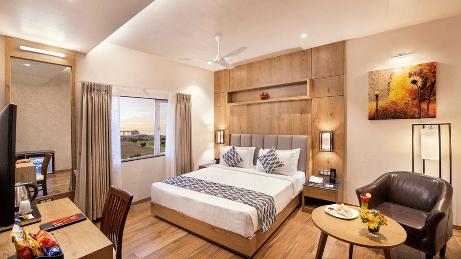 A hotel room in Lords Inn Jamnagar with modern interiors and wooden flooring featuring a double bed positioned near a window with a garden view, a mini bar, and a lounge chair
