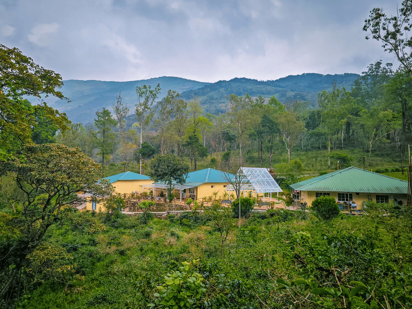 Ibex Resorts, Valparai (Mistly) buildings with green roofs located on a hillside, surrounded by trees, plants, pathways, and mountain ranges in the background under a cloudy sky.