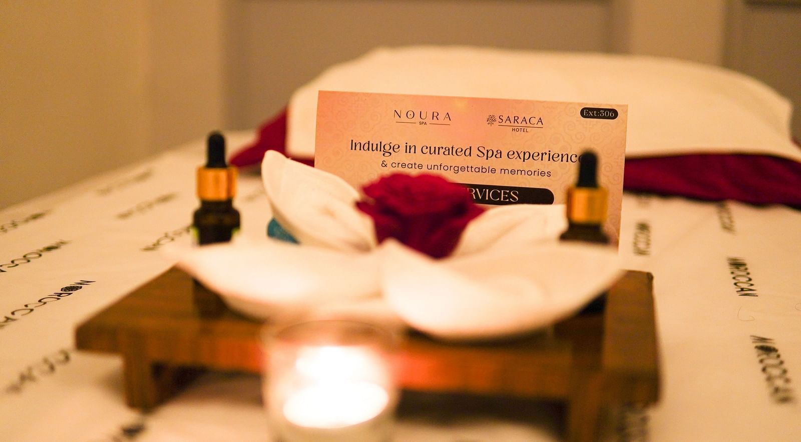 A card of Noura Spa placed on a massage bed along with essential oils and candle and a flower