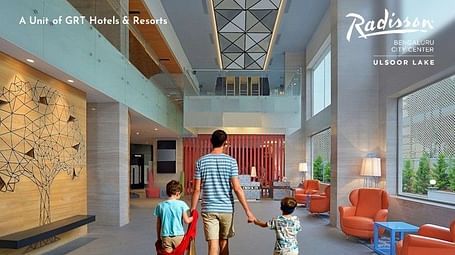 Creative family lifestyle photography of a father walking hand-in-hand with two young children through the hotel lobby, with one child carrying a red cape, showcasing the Independence Day Special family-friendly atmosphere at Radisson Bengaluru.
