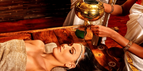 Person lying on a table receiving a stream of liquid poured onto the forehead by a practitioner using a suspended vessel during treatment