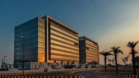 2 sleek, horizontal-banded office buildings reflecting a golden sunset next to a paved road lined with palm trees at Gurgaon.