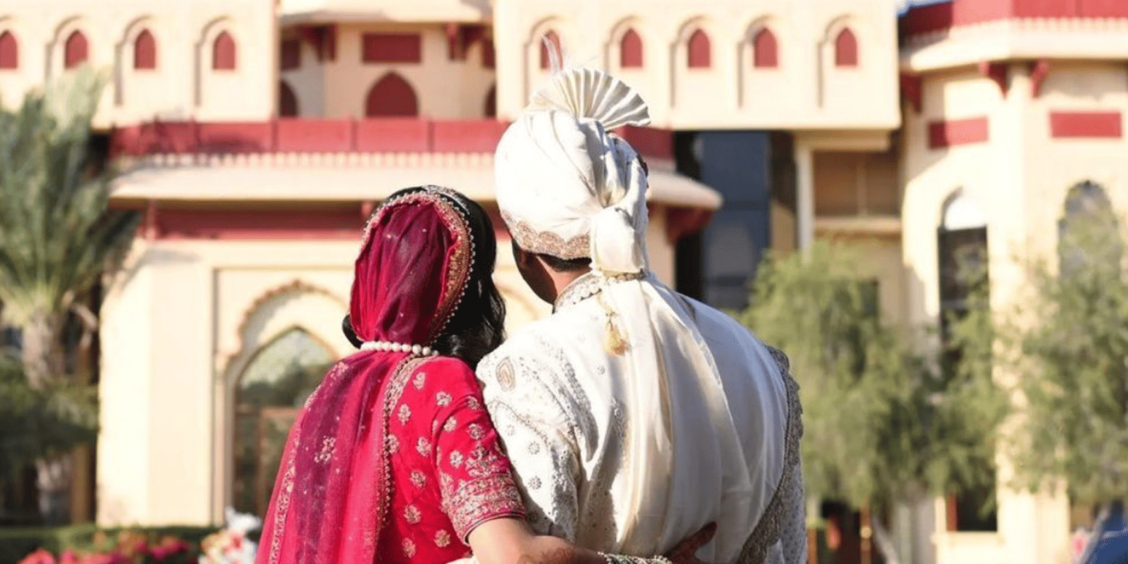 bride & groom in traditional Indian attire, standing in front of The Ummed Jodhpur Palace.