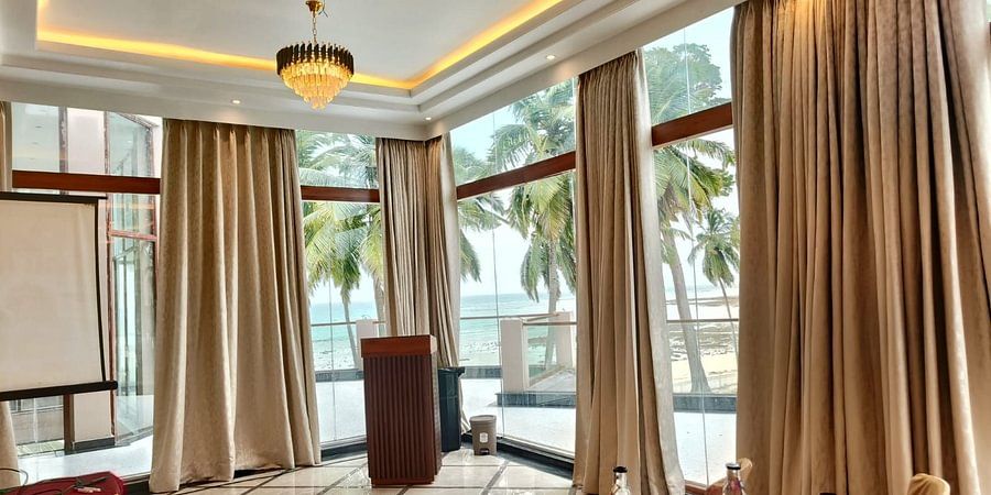alt-text An indoor banquet hall arranged for a formal event with beige and burgundy themed decor, round tables, chandeliers, and large windows offering a scenic view of palm trees