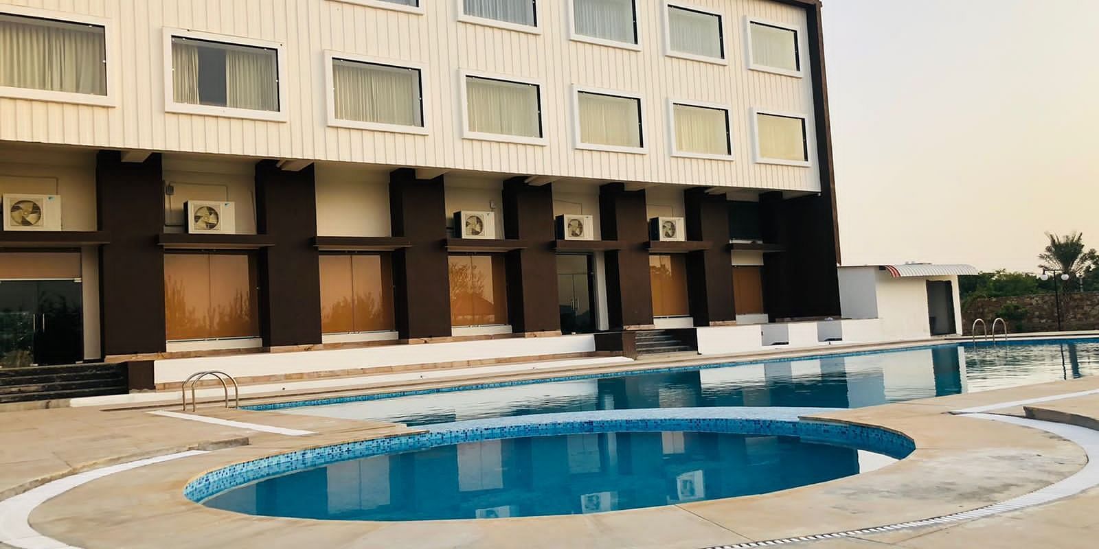 The exterior of the Bamboo Saa Mulberry Resort, Pushkar, featuring a multi-story building with a light-coloured facade with a swimming pool is visible in the foreground.