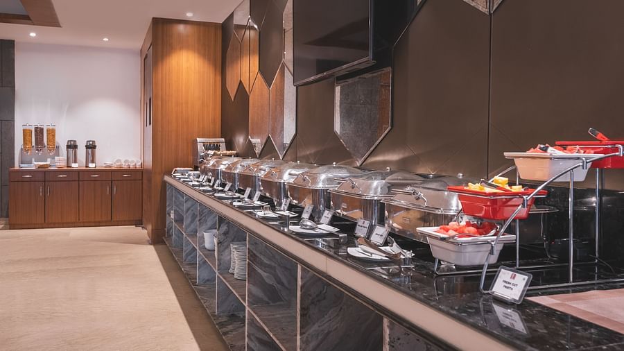 A well-stocked buffet line at Lords Inn Udaipur features covered chafing dishes and fresh fruit displays on a sleek black countertop.