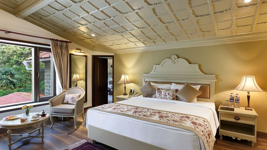 A full view of a room with a royal bed, chair, table and a huge glass window - the naini retreat, nainital.