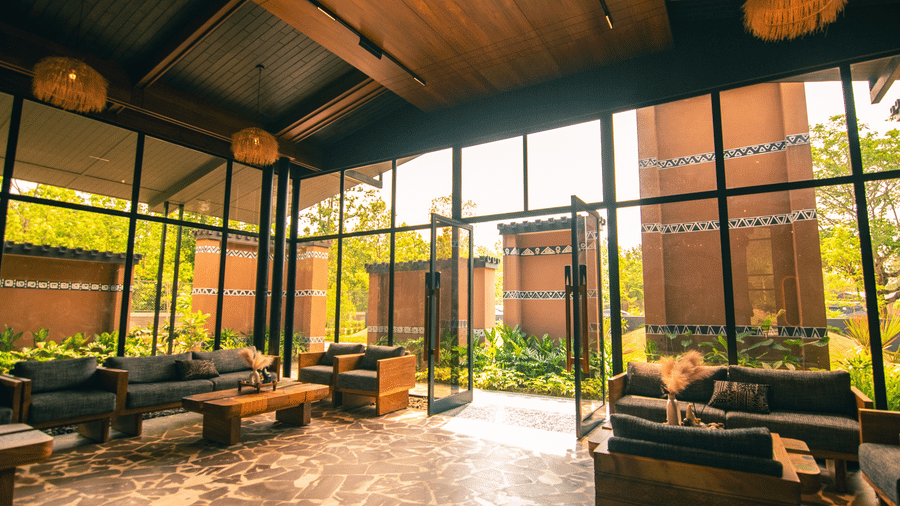 The lobby at Hotel Sonar Bangla Joypur Forest, Bankura features glass walls, cushioned seating with tables, and a door opening to a stunning view.