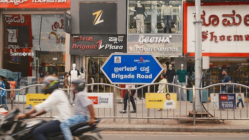 Street scene in Bangalore featuring shops, local signage in Kannada, and passing motorbike traffic.