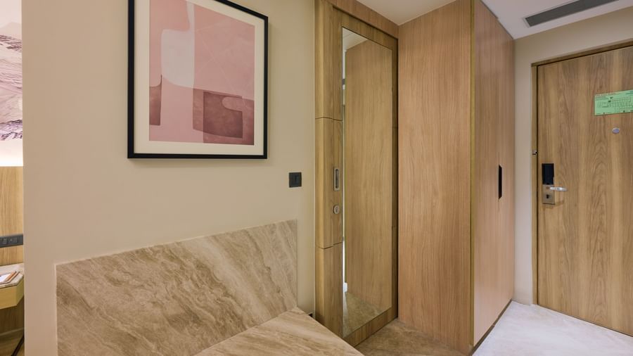 A hotel room foyer at Riviera Sarovar Portico, Pathankot, with a wooden wardrobe, mirror, and a small sitting bench.