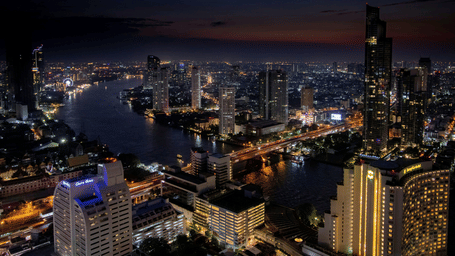 Aerial view of the Chao Phraya River in Bangkok at night, featuring illuminated skyscrapers, bridges, and city lights reflecting on the water.