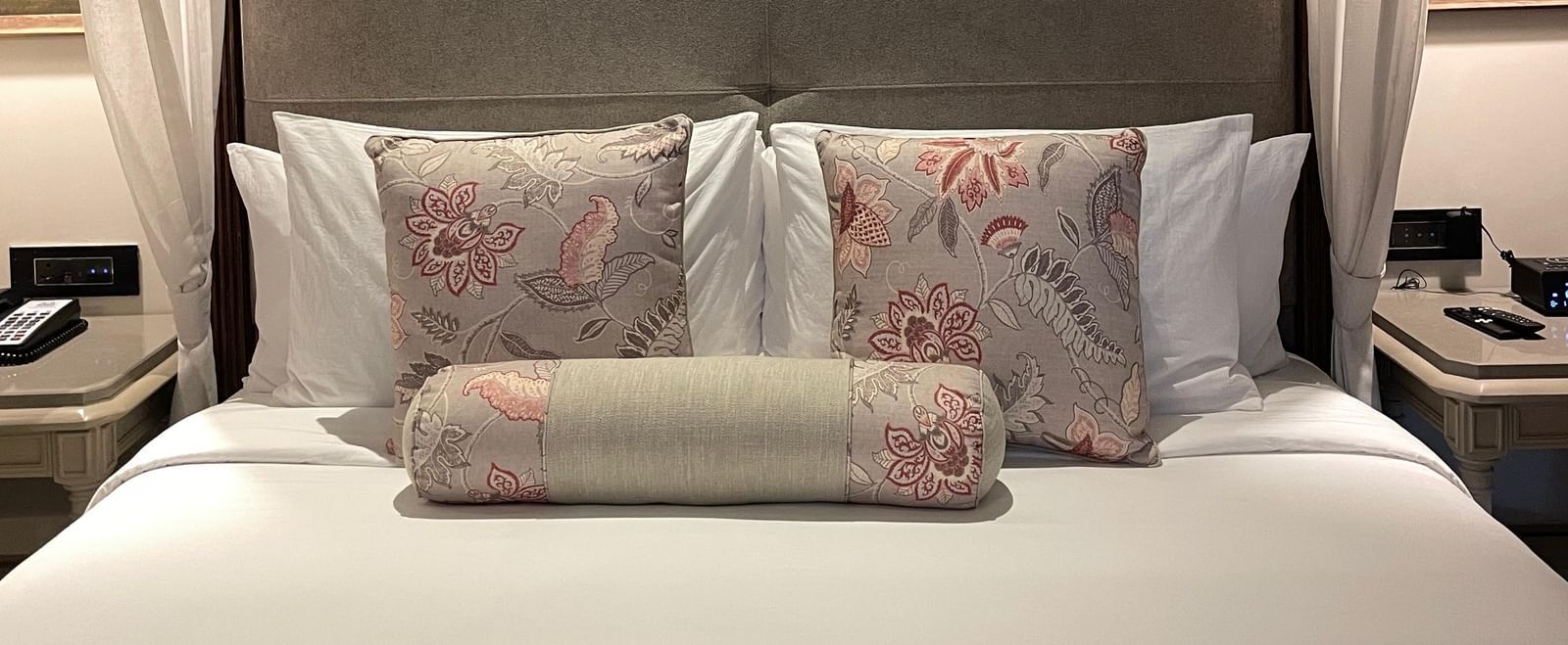 A close-up view of the king-sized four-poster bed in a Premium King Room at The Evren, Vagator, highlighting the neutral-toned headboard and the decorative pillows with floral patterns.