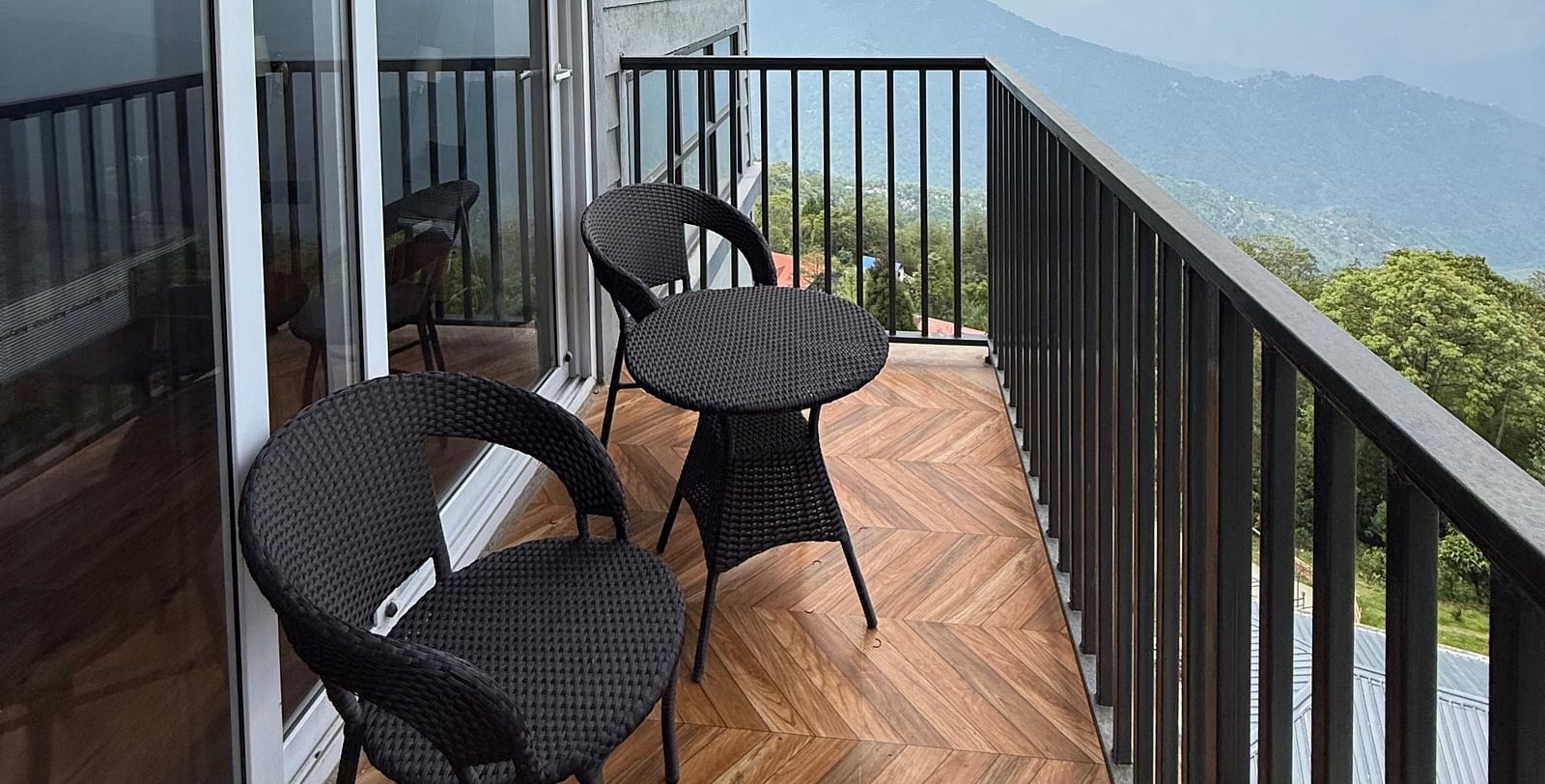 A modern balcony at Summit Enigma Resort & Spa, with black metal railings and herringbone-patterned flooring, offers a scenic view of distant mountains under a cloudy sky.   Sources