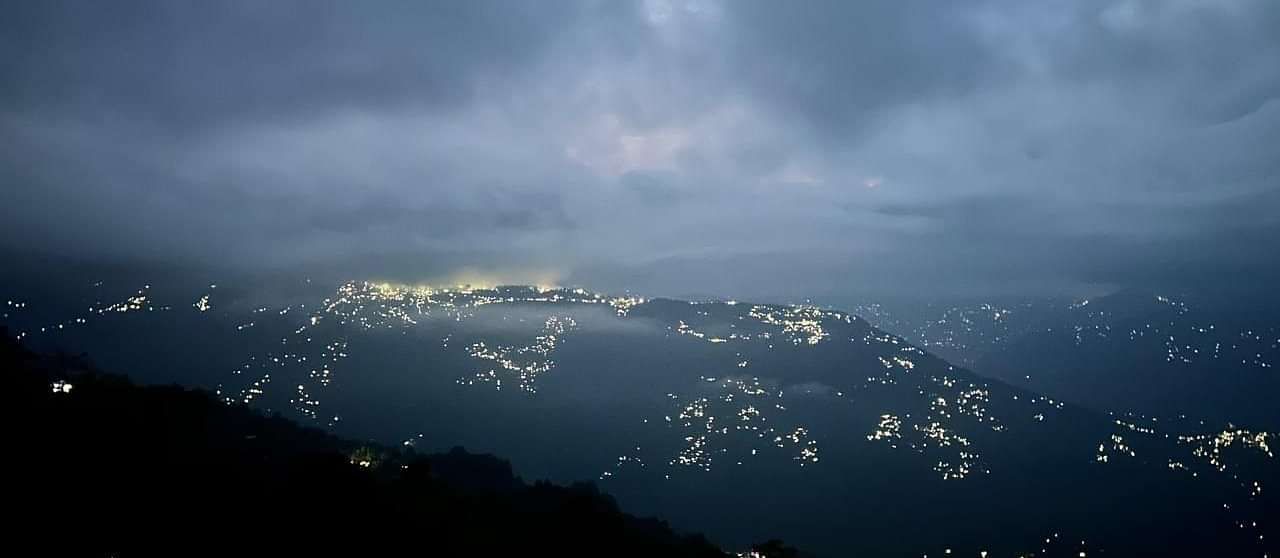  An evening view from Summit Enigma Resort & Spa shows a sprawling city twinkling with lights nestled in the hills under a dramatic cloudy sky.