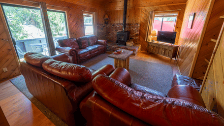A living room with wood-paneled walls and vaulted ceiling, featuring brown leather sofas, a brick fireplace with wood stove, wooden coffee table, TV unit, and large windows with tree views - Shaver Lake Village Hotel