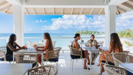 Guests dining by the sea under a shaded roof at Tamarind Hills Antigua – Resort & Villas, with a bright sky and ocean views
