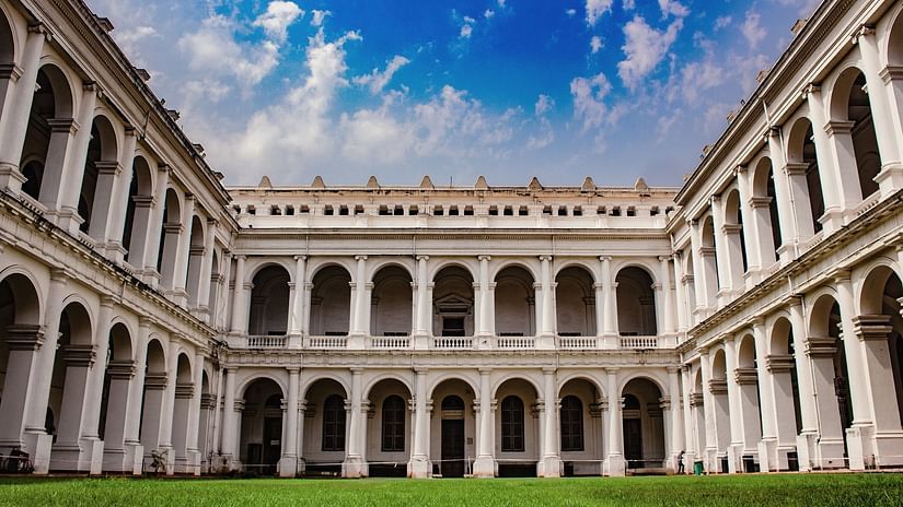 A grand white colonial-style building with arched corridors surrounding a green courtyard under a vibrant blue sky with scattered clouds.