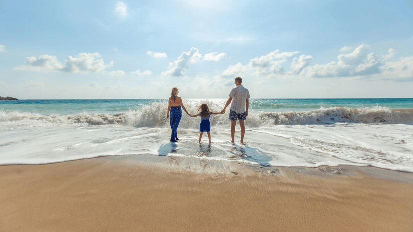 A family of three standing on the shore with waves brushing against their feet.