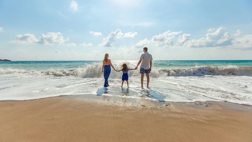 The image shows a family of three with two adults and a child, standing on a sandy beach, holding hands as waves gently reach them.
