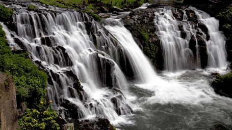 Multi-tiered waterfall cascading over dark rocky formations into a flowing river, surrounded by lush greenery and landscaped gardens.