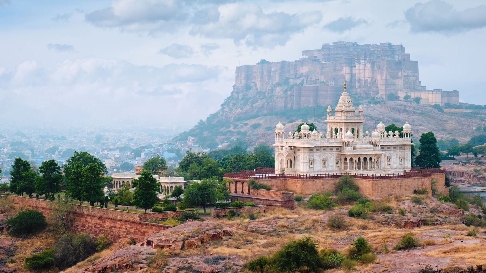 Distant view of Jaswant Thada, a white marble cenotaph in Jodhpur, set against the backdrop of Mehrangarh Fort and rocky terrain under a cloudy sky.