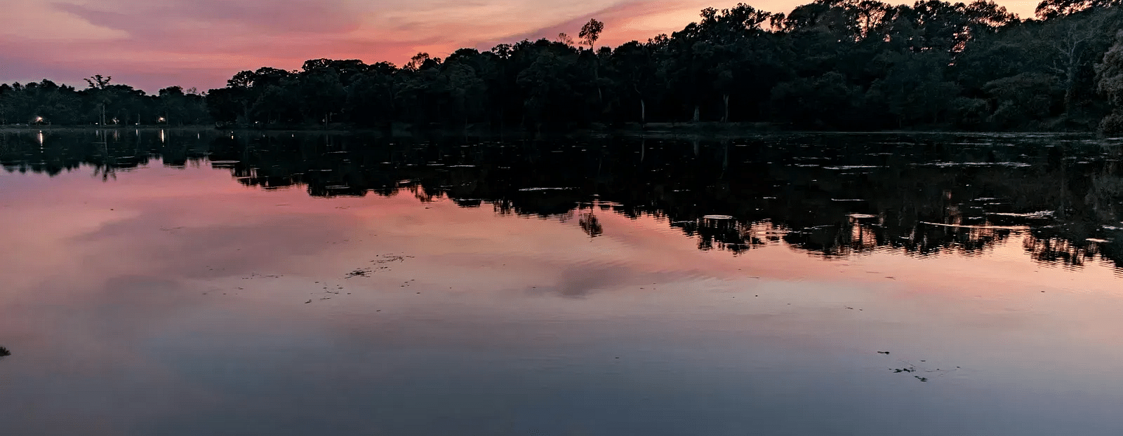 A serene lake at sunset with a colourful sky blending pink, purple, and orange hues, reflecting off the calm water surface, and a silhouette of trees lining the horizon.