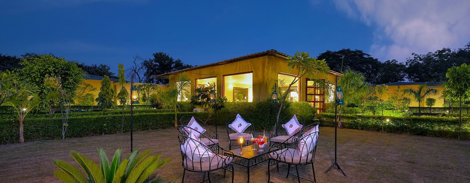 An outdoor dining setup at night featuring a table with four chairs, elegantly set with napkins and drinks, surrounded by greenery and illuminated by soft garden lighting, with a warmly lit building in the background under a deep blue sky - Singhvilas, Ran