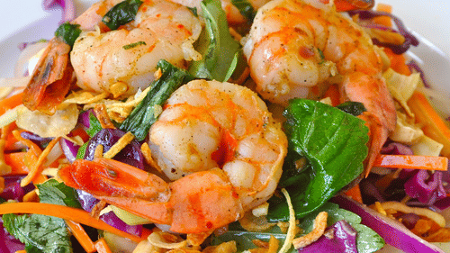 A plate of shrimp salad with vegetables including cabbage, carrots, and onions arranged on a white dish.