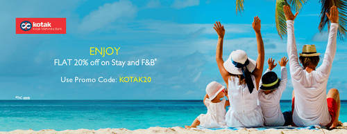 A cheerful family of four sits on a tropical beach facing the ocean, raising their hands in joy under a clear sky with palm trees. A promotional banner offers 20% off on stay and F&B for Kotak Mahindra Bank customers.