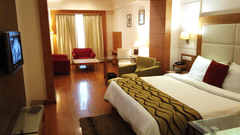 A Junior Suite at Ambrosia Sarovar Portico, Haridwar with a bed, seating area, a wall-mounted TV, wooden flooring, and curtains separating the living area.