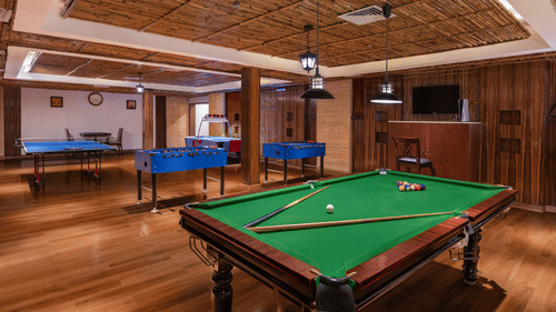 Spacious indoor recreation room featuring a snooker table, foosball tables, a ping pong table, and wooden flooring, with warm lighting and a cosy lounge ambience - Ananta Spa and Resort, Pushkar.