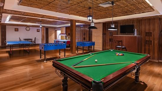 Spacious indoor recreation room featuring a snooker table, foosball tables, a ping pong table, and wooden flooring, with warm lighting and a cosy lounge ambience - Ananta Spa and Resort, Pushkar.