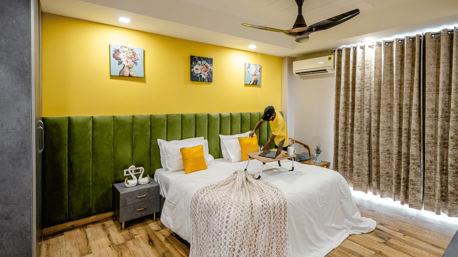 A housekeeping staff member in a yellow shirt making a double bed in a brightly lit hotel room with a green headboard.