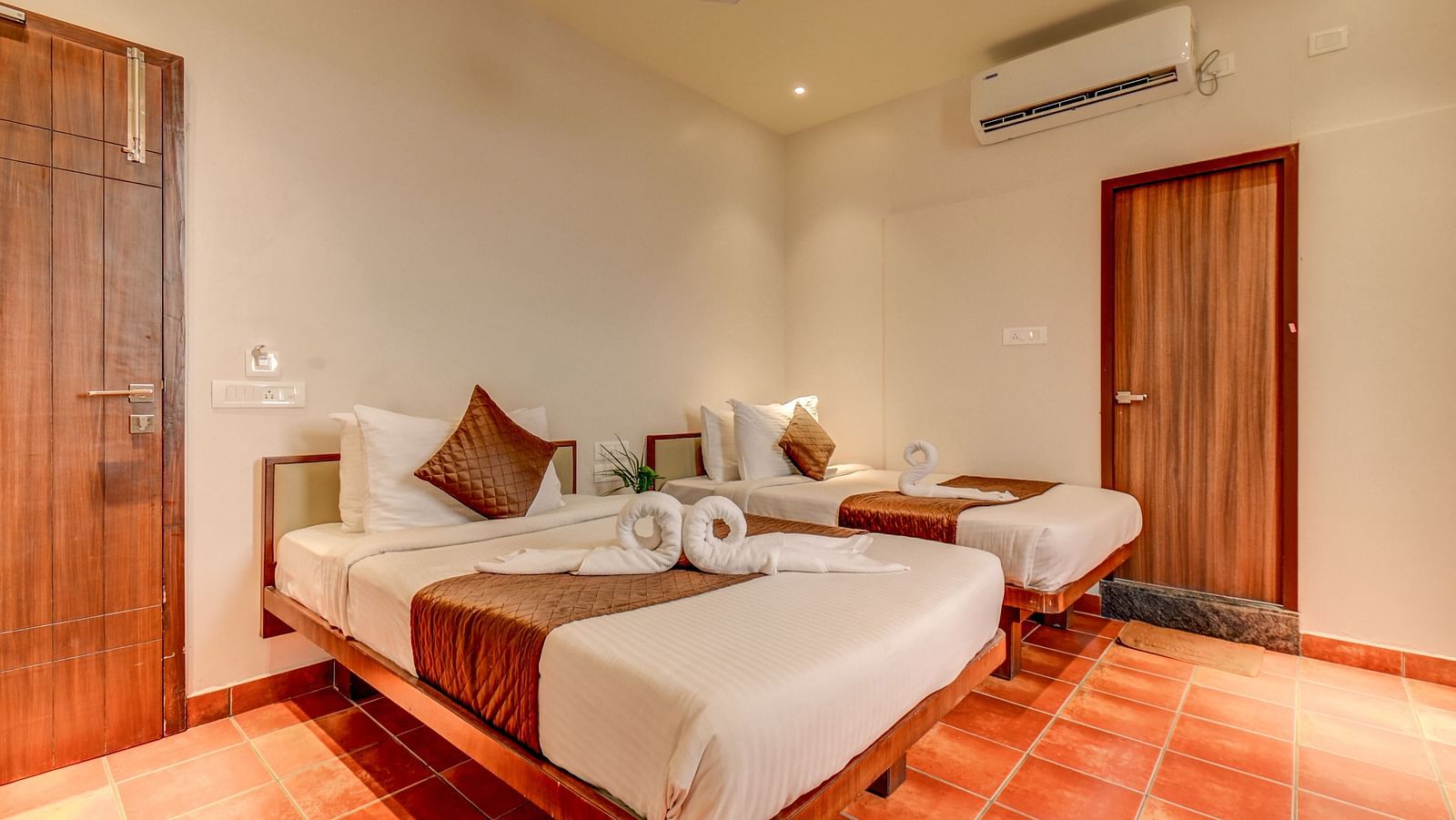 Two neatly made beds placed side by side, with doors on both sides and a wall-mounted AC above one door at Harigovinda's JNStay