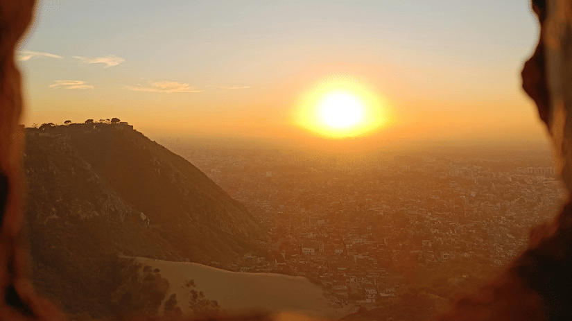 A bright sun sets over a hazy horizon, captured through a small, rectangular stone window opening in a fort wall.