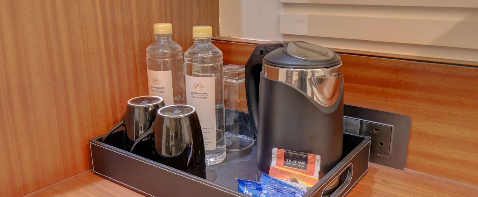 An image of a flat surface with a tray containing two water bottles, a kettle, two cups, and packets of milk and tea at Hari Govindas, Siddappa Square.