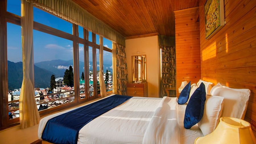 An image of a room with a neatly arranged bed with the view of a mountain city through glass windows, complemented by wooden interiors - Central Heritage Resort and Spa,  The Mall Road, Darjeeling