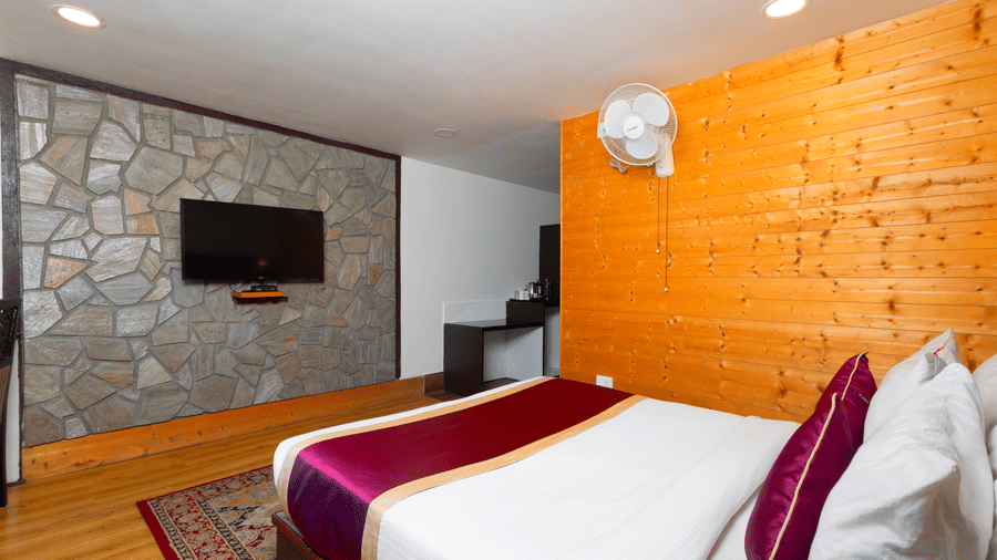 Modern deluxe room at Sumitel Hotels featuring a king-size bed with white linens and purple accents, wooden wall panels, warm lighting, and a cozy, elegant mountain stay ambiance.