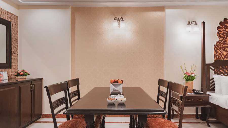 An organised dining space in a well lit room with pendant lights featuring a partially visible bed, a bedside table, a cabinet and a wall mirror at Shri Vallabh Vilas Lords Plaza Nathedwara.