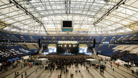 High-angle view of a stage and large crowd inside a massive indoor stadium with a complex white roof structure.