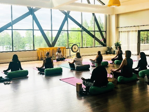 A group of people are meditating on cushions and mats in a spacious room with large windows and a high ceiling at YO1 Longevity & Health Resorts
