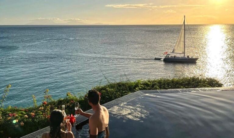 A couple enjoying drinks in an infinity pool overlooking the ocean at sunset, with a sailboat gliding across the water in the distance.