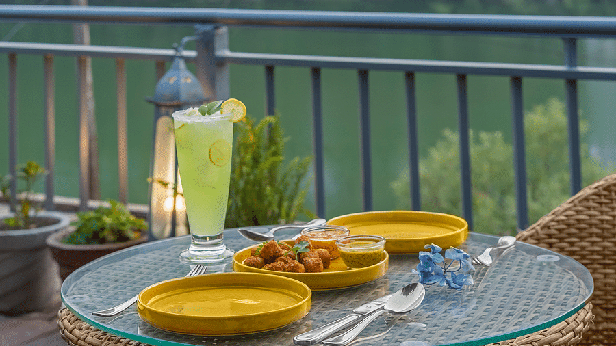 A scenic outdoor dining area with a glass table and round plate and a glass of cooling drink, overlooking a tranquil lake. lantern and greenery create a relaxing atmosphere.